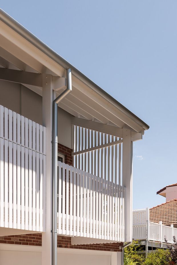 Close-up exterior view of a light-painted residential structure with a pitched roof, exposed eaves, gutters and downpipes, vertical slatted screening, and a covered balcony or verandah. Red brick walls and neighbouring tiled-roof homes are partially visible. The image is bright and modern, with strong linear detailing and a suburban residential context.