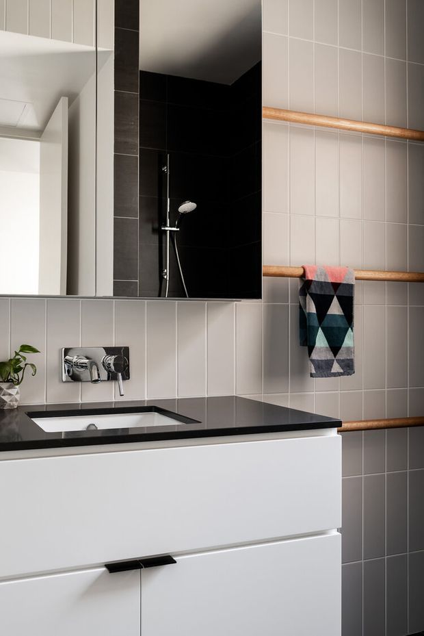A contemporary bathroom vanity with a white wall-mounted drawer unit, black stone benchtop, integrated white basin, and chrome wall-mounted tapware. Large-format grey tiled walls create a clean backdrop, with a black tiled shower recess and frameless mirror. Timber towel rails and a geometric multicoloured towel add warmth and contrast.