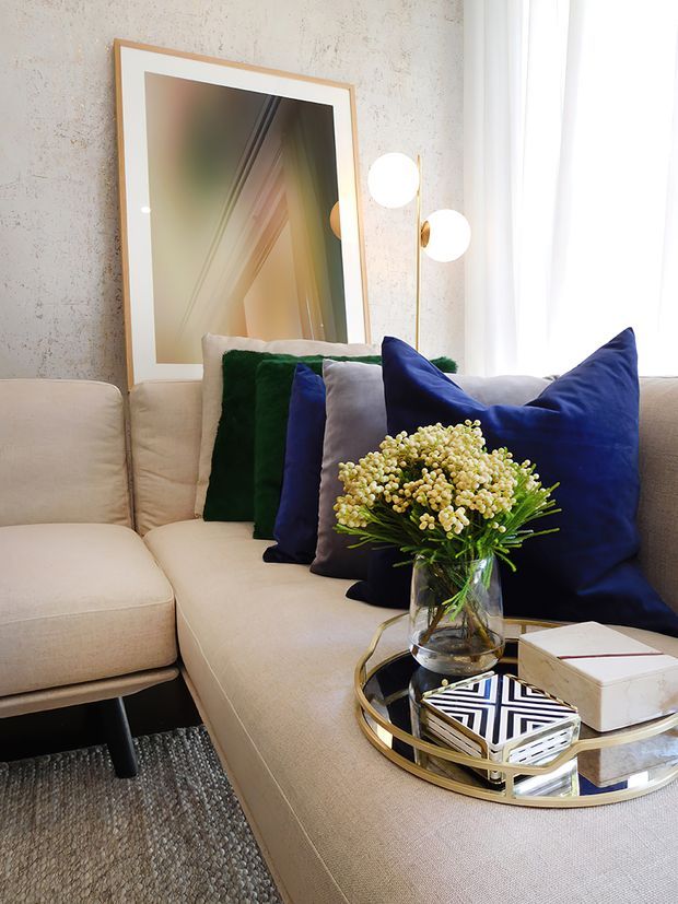 A close-up of a refined living room seating area with a large cream upholstered sectional sofa, deep green, navy and grey cushions, and a round mirrored coffee table with a gold-toned frame. A glass vase of flowers, books and decorative objects sit on the table. The room has a textured pale wall, a large framed mirror or artwork, and a sculptural brass-and-white globe floor lamp beside sheer curtains. The overall look is polished, comfortable and colourfully accented.