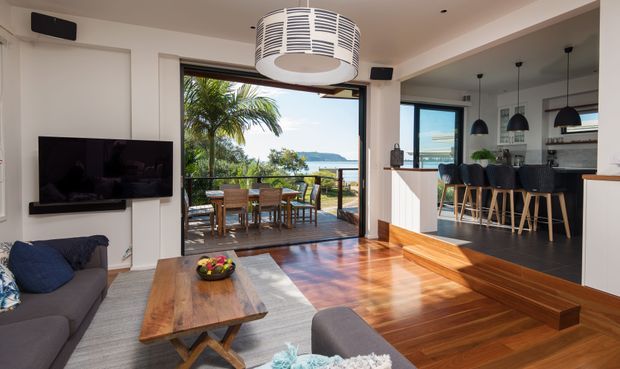 Bright open-plan living and kitchen interior opening onto a timber deck with an outdoor dining area and coastal water views. The room features polished timber flooring, white walls and cabinetry, a raised kitchen platform with a dark floor, pendant lights over a breakfast bar, a wall-mounted television, and contemporary furniture. A large sliding glazed opening provides strong indoor-outdoor connection.