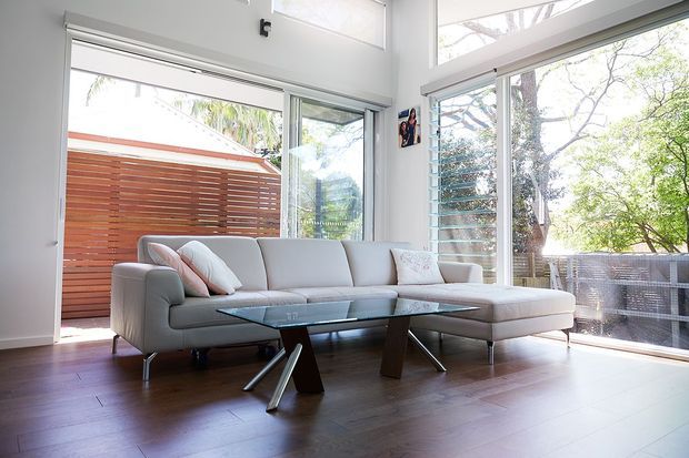 Bright contemporary living room with a large light-grey sectional sofa, glass coffee table, dark timber flooring, white walls, and extensive floor-to-ceiling glazing. The open doors connect to a timber privacy-screened outdoor area, while louvred glazing and trees are visible beyond.
