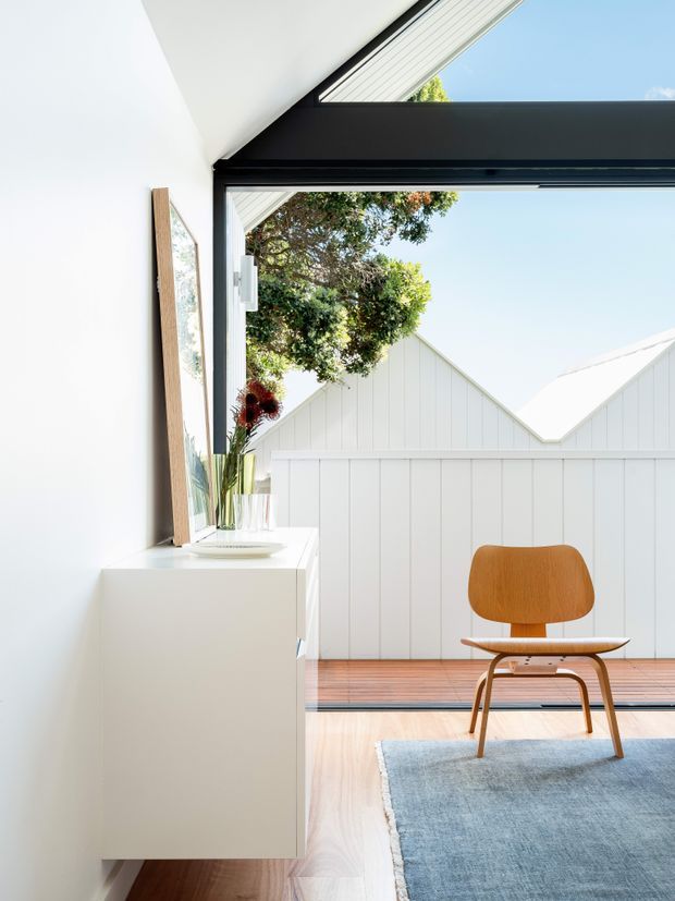 Bright minimalist interior corner with a white built-in sideboard, framed artwork and vase, opening onto a timber-floored terrace or balcony. A warm mid-century style wooden lounge chair sits on a pale blue rug beside large black-framed glazing. Outside, a white vertical-panel gabled structure and leafy tree are visible under a clear blue sky.