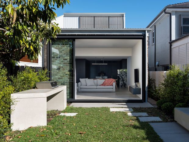 Rear exterior view of a contemporary residential extension opening onto a landscaped courtyard. The addition has a broad black-framed glazed opening, white rendered walls, a projecting flat roof, and an upper level with vertical metal screening. Inside, a living room with a light sectional sofa and dark built-in cabinetry is visible. A white built-in outdoor bench or counter sits in the foreground beside stepping-stone paving and planting.