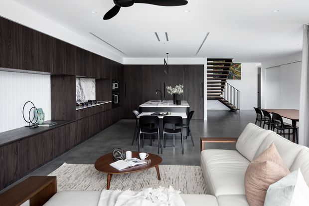 Contemporary open-plan interior combining a living room, dining area and kitchen. Dark timber cabinetry lines the left wall, with a pale stone island and dining table at the centre. A white sectional sofa, timber coffee table and textured rug occupy the foreground, while a staircase with open timber treads is visible beyond. The space has a clean, refined monochromatic palette with warm wood accents and recessed lighting.
