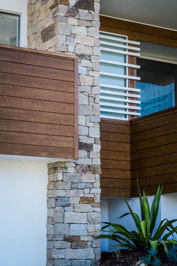 Close-up exterior architectural detail showing a modern facade with a prominent rough-faced stacked stone column, dark timber horizontal cladding and balcony privacy screens, white-framed glazing with horizontal timber sun-shading battens, and landscaped planting at the base. The composition highlights material contrast and projecting architectural elements.
