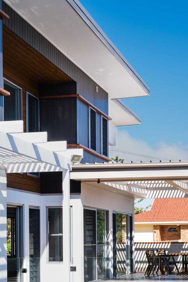 A bright exterior view of a contemporary multi-level residential house, focused on the facade and covered outdoor terrace. The building features crisp white rendered walls, dark vertical cladding, warm timber soffits and trims, extensive dark-framed glazing, projecting flat roof planes, and a slatted pergola casting strong linear shadows over the tiled patio and dining area. A neighbouring tiled roof is visible in the background.