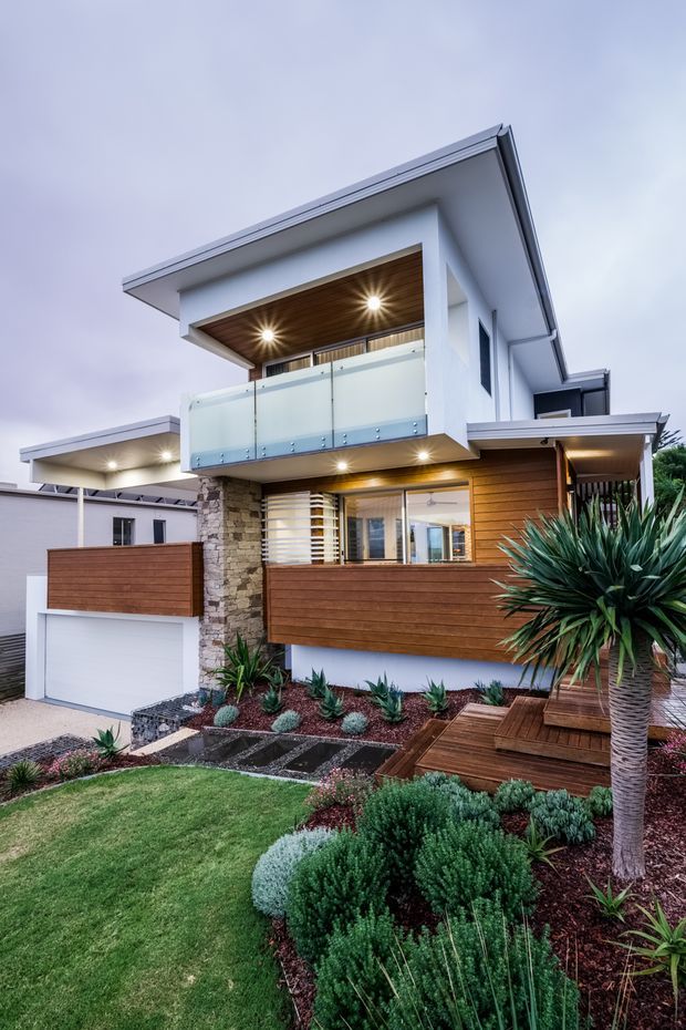 Exterior view of a contemporary multi-level home with crisp white rendered walls, broad flat roof overhangs, warm horizontal timber cladding, a stone-clad feature column, and a frosted-glass balcony. The landscaped front garden includes lawn, low sculptural planting, dark gravel, stepping stones, and a timber deck beside the garage and entry.