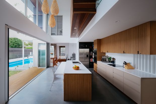 Bright contemporary kitchen in a high-ceilinged open-plan space, featuring a large central timber island, extensive white cabinetry, a white tiled splashback with vertical fluted detailing, dark flooring, and integrated stainless-steel appliances. Floor-to-ceiling glazing opens toward a timber deck and swimming pool, while a timber-lined ceiling feature and woven pendant lights add warmth.