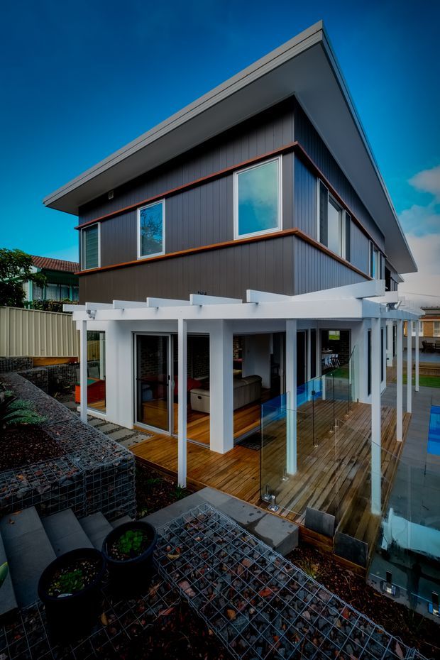 Exterior view of a contemporary multi-level house with a sharply projecting flat roof, dark vertical cladding, white structural columns and beams, extensive glazing, timber decking, and a swimming pool beside the lower-level outdoor living area. The elevated camera angle also shows gabion retaining walls and landscaped terraces.
