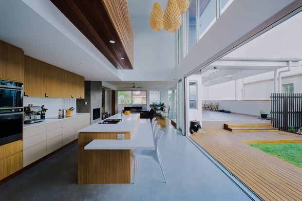 Bright open-plan contemporary kitchen and living space with a long timber-and-white island, integrated cabinetry, concrete-look flooring, sculptural pendant lights, and floor-to-ceiling glazing opening onto a timber deck and courtyard. The room has a double-height glazed connection to the outdoor area and a clean, refined material palette.