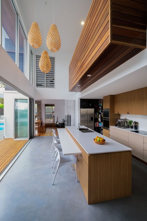 A bright, contemporary open-plan kitchen with a long white island and integrated dining overhang, warm vertical-grain timber cabinetry, white tiled splashback, stainless-steel appliances and a smooth grey concrete floor. The space has a high ceiling with three woven pendant lights, extensive glazing and an open connection to a timber deck and adjoining living area. A large timber-clad overhead volume adds architectural definition.