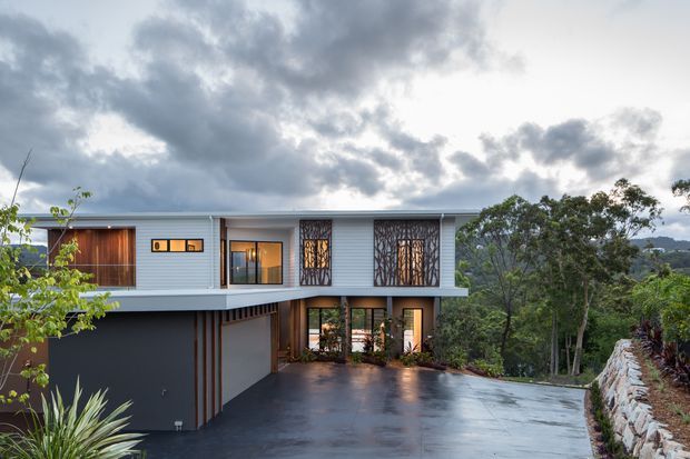 Exterior view of a contemporary elevated two-storey residence set into a sloping, wooded site. The house has pale horizontal cladding, dark charcoal lower walls, timber-clad entry elements, large glazed openings, decorative dark metal screens, and a broad flat roofline. A wet paved driveway and stone retaining wall occupy the foreground, with dense vegetation and distant hills beyond.