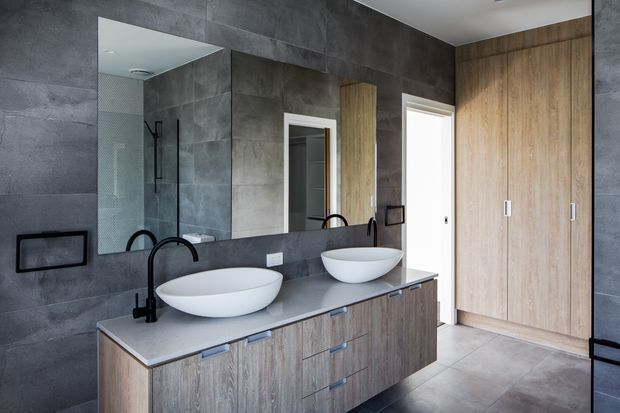 Contemporary double-vanity bathroom with a long grey stone benchtop, two white vessel basins, matte black tapware, dark grey wall and floor tiles, and extensive timber-look built-in cabinetry. A large mirror, glass shower enclosure, and full-height wardrobe-style storage create a clean, refined fit-out.