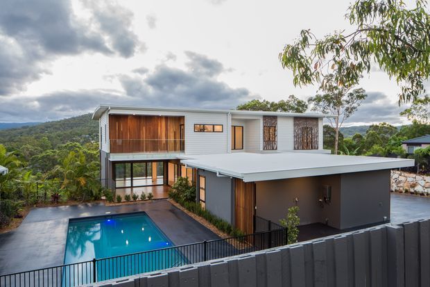 Exterior view of a contemporary multi-level residence on a landscaped, sloping site. The house features a broad low-pitched roof, clean horizontal forms, grey rendered walls, light grey horizontal cladding, warm timber screening and cladding, expansive glazing, and a private rectangular swimming pool in the foreground. Dense greenery and rolling hills surround the property.