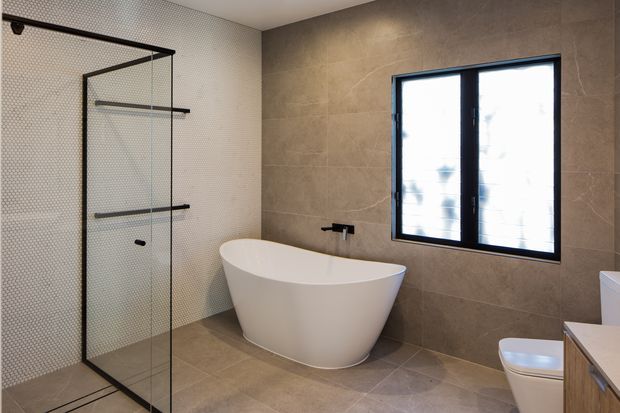 Contemporary bathroom featuring a freestanding white oval bathtub, frameless glass shower enclosure with black hardware, large beige stone-look wall and floor tiles, and a black-framed window. A vanity and toilet are partially visible at the right edge.