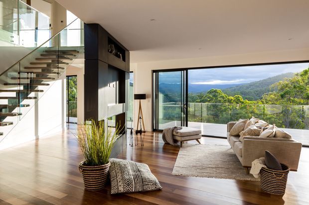 Bright contemporary open-plan living room with polished timber flooring, a large neutral sectional sofa, a textured area rug, and floor-to-ceiling glazing opening onto a balcony with expansive green hillside views. A sculptural floating staircase with glass balustrade and dark built-in media cabinetry define the space.