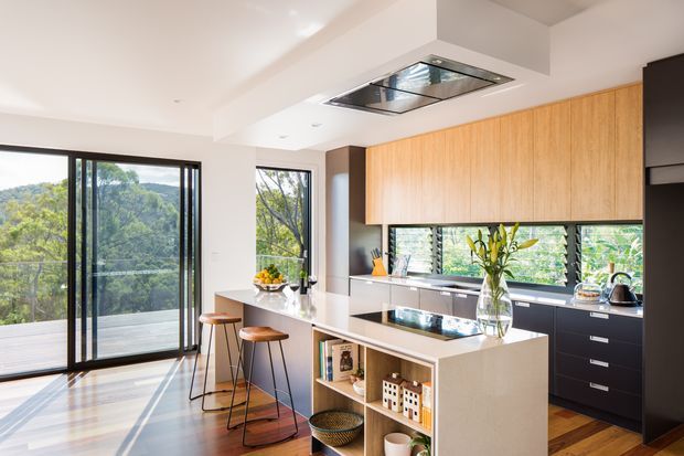 Bright contemporary kitchen with a large central island, integrated cooktop, white stone surfaces, warm timber overhead cabinetry, and dark lower cabinetry. Full-height glazed sliding doors open to a balcony or terrace with leafy views, while a horizontal louvred window brings additional natural light above the benchtop. The room has polished timber flooring, a white ceiling with recessed lighting, and a skylight or roof window above the island.