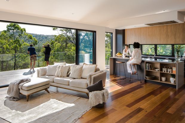 Open-plan contemporary living and kitchen area with expansive floor-to-ceiling glazing opening to a balcony overlooking a leafy wooded landscape. The room features a large light-coloured sectional sofa, pale area rug, timber flooring, a dark kitchen island with bar seating, and warm timber cabinetry extending across the kitchen wall. Abundant natural light creates an airy, inviting atmosphere.