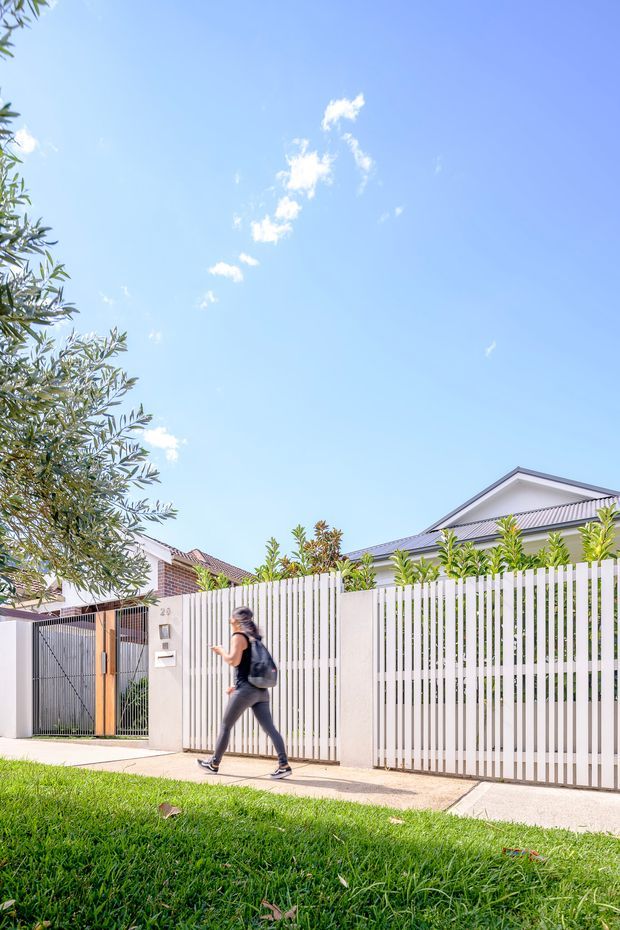 Bright daytime streetscape showing a contemporary residential property frontage with a white vertical-slat privacy fence and matching sliding gate, pale rendered masonry walls, a corrugated pitched roof, and a pedestrian sidewalk beside a grass verge. A neighbouring brick-roofed home, established planting, and a walking person are also visible.