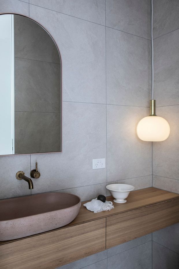 Contemporary bathroom vanity with large grey stone-look wall tiles, a rounded rectangular arch-top mirror, a pinkish stone vessel basin, brushed brass wall-mounted tapware, and a floating timber vanity with integrated storage. A warm globe pendant adds a soft accent above the timber benchtop.