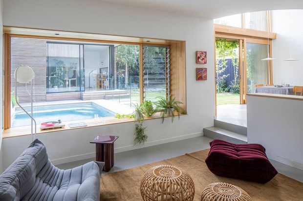 Bright contemporary living room opening to a landscaped courtyard and swimming pool. The room features expansive timber-framed glazing, a grey upholstered sofa, sculptural burgundy ottoman, woven round coffee tables, indoor plants, and a partial kitchen or dining area beyond a raised step. The large window provides a strong visual connection to the pool and outdoor space.