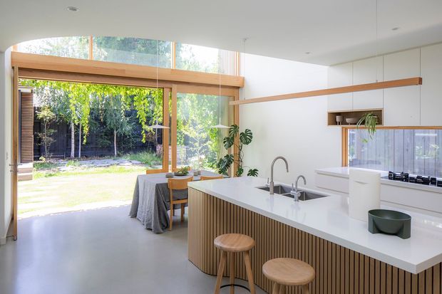 Bright contemporary kitchen with a large white island featuring fluted timber-panel cabinetry, integrated sink and curved faucet, and two timber stools. A dining table sits beside extensive floor-to-ceiling glazing that opens toward a garden. White walls, pale polished flooring, timber-framed glazing, simple linear pendant lights, and minimal cabinetry create an airy, refined interior.