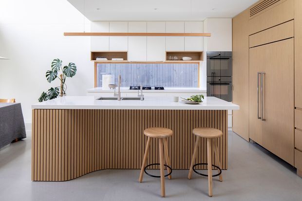 Bright contemporary kitchen with a large white island featuring a curved slatted timber front, two timber stools, integrated sink and cooktop, pale cabinetry, timber wall storage, and a vertically ribbed blue-grey splashback. A linear timber pendant and indoor plant add warmth to the clean, open space.