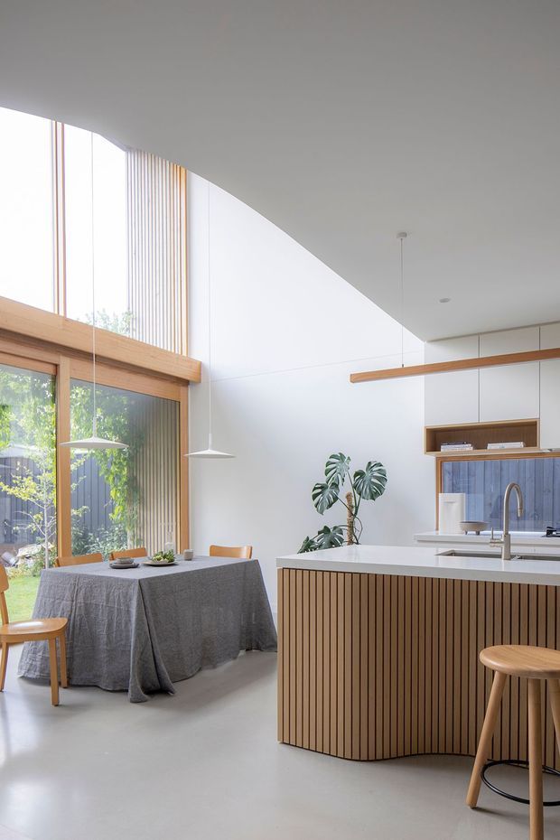 A bright, open-plan kitchen and dining area with a sculptural curved ceiling and full-height glazing. The kitchen island has a white benchtop and vertically slatted timber-clad base with a curved end, paired with timber stools. A compact dining table with a grey textile tablecloth sits beside large timber-framed windows. White cabinetry, a blue-grey tiled splashback, pendant lights, and indoor greenery create a clean, contemporary atmosphere.