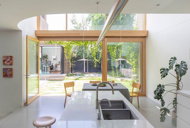 Bright contemporary kitchen centered on a large white island with an integrated double-bowl stainless-steel sink and curved chrome tap. Warm timber-framed floor-to-ceiling glazing and a high clerestory-style window open directly to a leafy courtyard, with a dining table and chairs beyond. White walls and flooring create an airy, indoor-outdoor setting, complemented by a large indoor plant and colourful wall art.