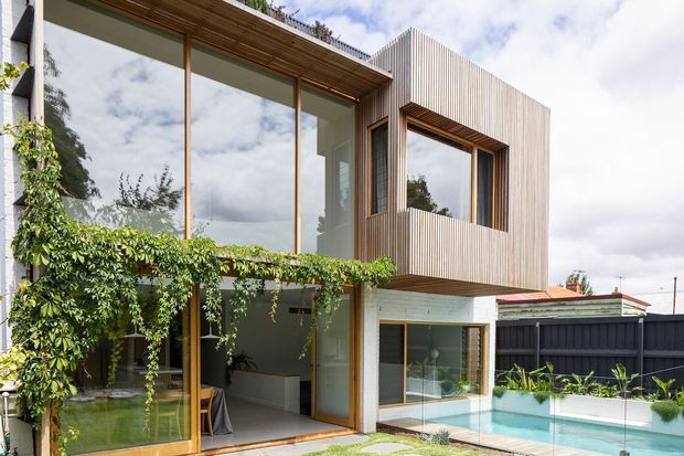 Contemporary two-storey residential facade viewed from a landscaped courtyard, with extensive floor-to-ceiling glazing, warm timber window and door frames, vertical timber cladding to the upper level, and a sheltered ground-floor living area opening toward a swimming pool. Climbing greenery softens the facade, while a dark boundary fence and neighbouring roof are visible in the background.