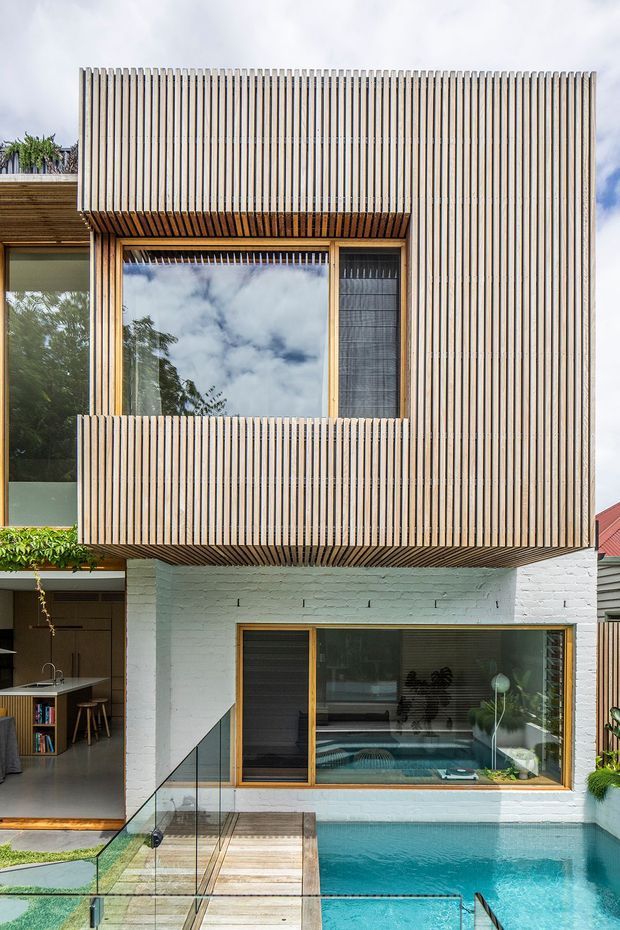 A contemporary multi-level residential facade with a white-painted brick lower storey and a projecting upper volume clad in closely spaced vertical timber battens. Large timber-framed windows open toward a compact turquoise swimming pool, with a glass balustrade and timber deck in the foreground. An open kitchen area is visible at the left edge, while reflected trees and sky appear in the glazing.
