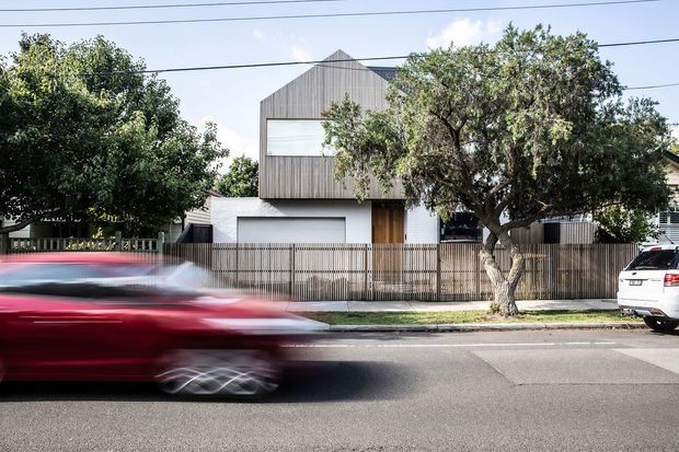 Street-facing view of a contemporary multi-level residence with a steep gable-front form, vertically clad upper storey, white rendered lower level, timber entry door, and a narrow vertical-slat front fence. Mature street trees and motion-blurred traffic occupy the foreground.