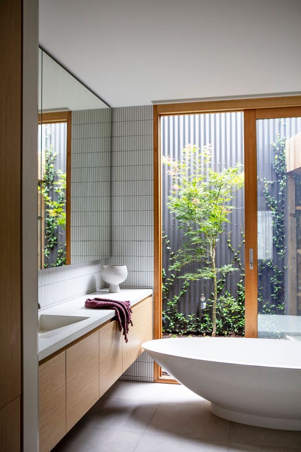 Bright contemporary bathroom featuring a freestanding white bathtub beside full-height timber-framed glazing, a narrow planted courtyard, a long light-wood vanity with integrated basin, large mirror, and vertically gridded pale tile wall finish. The room has a calm, refined, natural palette and strong indoor-outdoor connection.