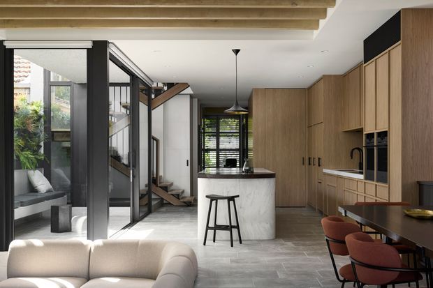 Open-plan contemporary kitchen, dining and living area with extensive warm timber cabinetry, a curved stone island, integrated appliances, dark-framed internal glazing, exposed timber ceiling beams and a dining table with upholstered chairs. A stairway and glazed courtyard connection are visible beyond the kitchen.
