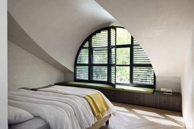 A serene bedroom with a low-profile bed dressed in white bedding and a mustard-yellow throw. The room features a dramatic arched window with black-framed glazing and horizontal louvres, an integrated dark timber window seat and low storage joinery, pale walls, and light timber flooring. Strong sunlight filters through the louvres, creating striped shadows across the floor.
