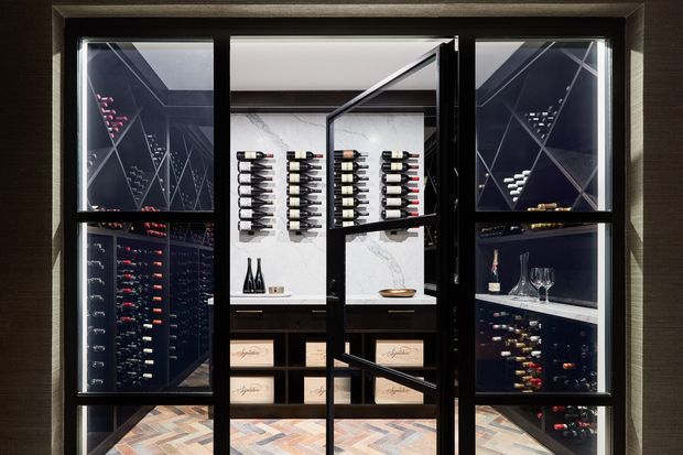 A compact, dark-toned wine storage room viewed through a black-framed glazed enclosure. Floor-to-ceiling wine racks line the side walls, while the rear wall features display racks, bottles, drawers, and a light marble-look backing. Geometric black framing and reflective glazing create a refined, gallery-like presentation, with patterned timber flooring visible at the base.