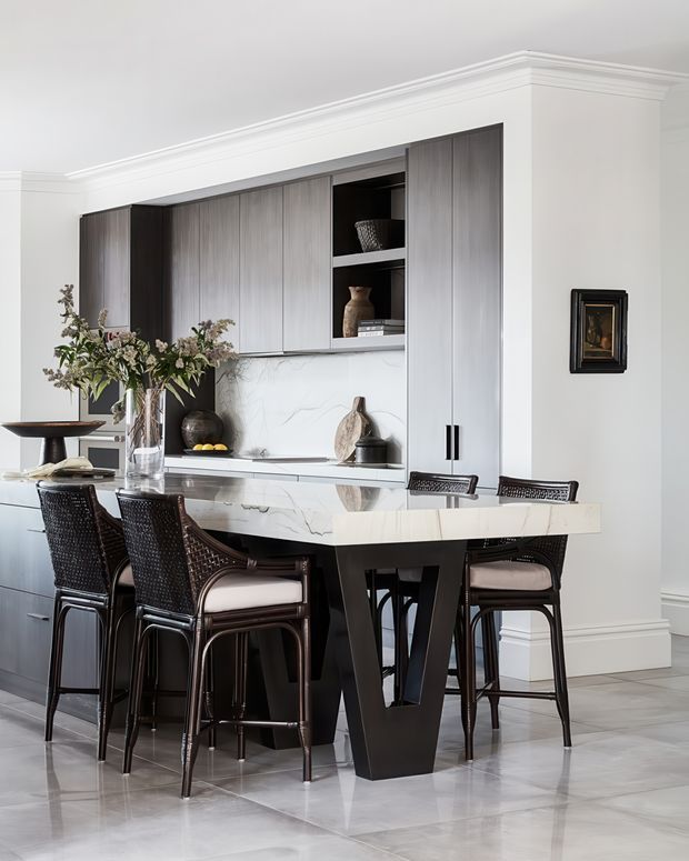 A refined open-plan kitchen and dining area with a large marble-look island, dark integrated cabinetry, and four woven bar stools. The room features white walls with substantial traditional crown moulding and skirting, pale large-format flooring, a minimalist white splashback, black accents, and decorative open shelving. Soft natural light and a restrained neutral palette create an elegant, polished atmosphere.