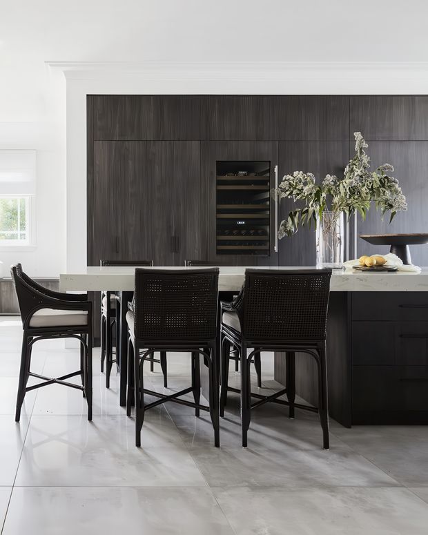 A refined contemporary kitchen and dining area with a long white stone island, dark wood-grain full-height cabinetry, integrated wine storage, and black woven bar stools. Large pale stone-format floor tiles, white walls with classic cornice detailing, and sculptural floral arrangements create a bright, high-contrast interior.