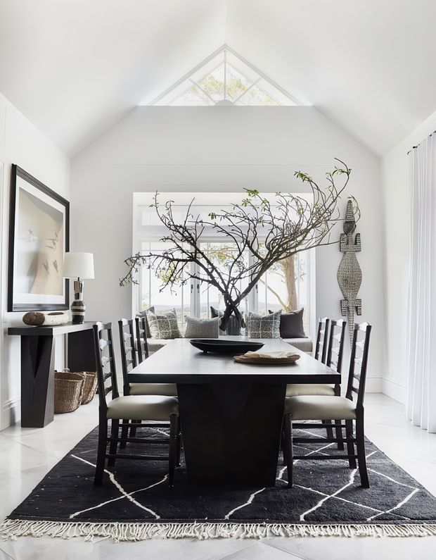Bright, refined dining room with a long dark rectangular dining table and matching dark chairs arranged on a black geometric rug. White walls, a vaulted ceiling with a triangular clerestory window, expansive glazing, sheer white curtains, and a sculptural branch arrangement create an airy monochrome interior. A console, artwork, baskets, and decorative wall sculpture provide secondary styling.