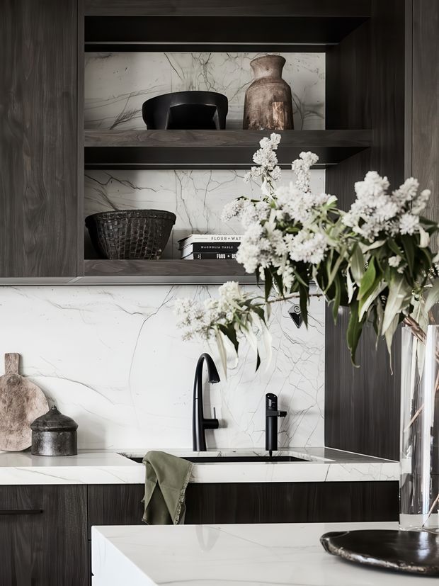 Close-up view of a refined contemporary kitchen featuring dark vertically grained cabinetry, open display shelving, a white marble-look splashback and benchtops with prominent grey veining, and matte black sink fixtures. Decorative vessels, books, greenery, and white flowers add a styled, luxurious character. A kitchen island or peninsula is visible in the foreground.