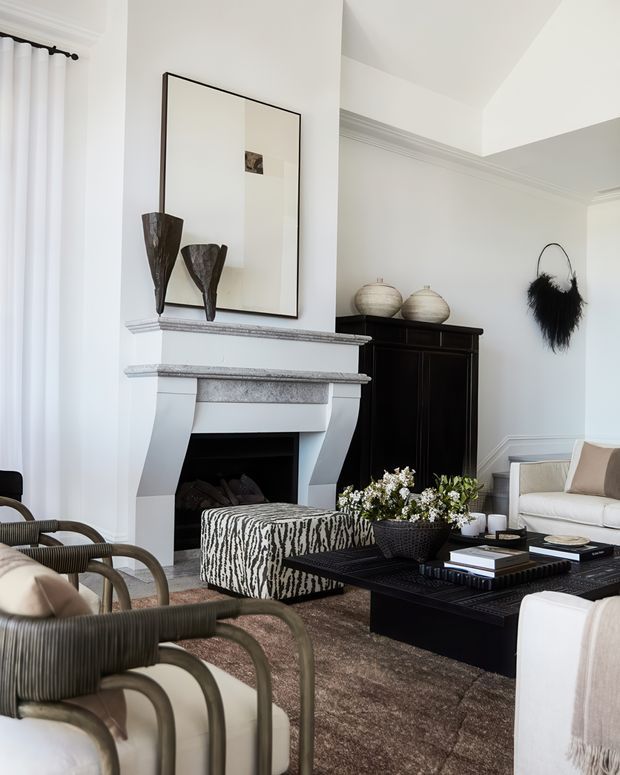 Elegant, bright living room with a white sculptural fireplace mantel, large framed mirror, dark timber cabinet, black coffee table, neutral upholstered seating, woven armchairs, and a zebra-patterned ottoman. The room has high ceilings, decorative ceiling mouldings, white walls, and a refined monochromatic palette with warm brown flooring and textiles.