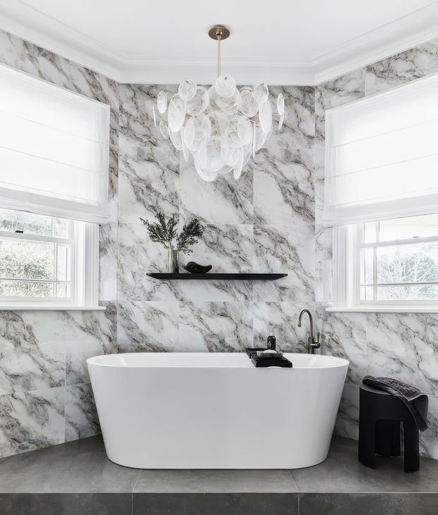 A bright, refined bathroom centered on a freestanding white bathtub. The walls are fully clad in dramatic white-and-grey marble-look panels, complemented by dark grey floor tiles, white plantation-style window shades, black tapware, and a slim black floating shelf. A sculptural white pendant light hangs above the bath, while a small black stool with a folded textile sits to the side.