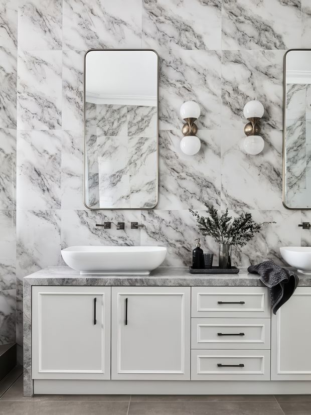 A refined contemporary bathroom vanity area with a large double-basin-style arrangement, white vessel basins, white paneled cabinetry, a grey-and-white marble-look tiled wall and benchtop, rounded rectangular mirrors, and globe wall sconces. Dark hardware, accessories, and a folded towel provide contrast against the predominantly monochromatic scheme.