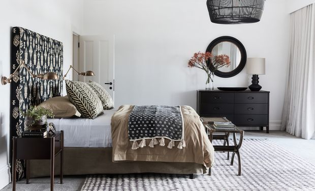 A refined bedroom with a low upholstered bed, layered neutral bedding, and a bold black-and-white patterned upholstered headboard and throw. A dark timber dresser with a round black-framed mirror, bedside table, lamps, books, and a glass-topped bench complete the room. White walls, full-height curtains, patterned rugs, and a sculptural pendant create a polished, monochromatic interior.