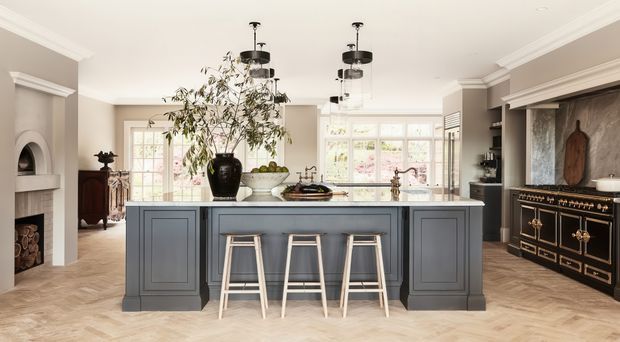 A spacious, refined traditional kitchen centred on a large dark grey island with paneled ends and three light timber stools. The room features a herringbone timber floor, white coffered or detailed ceiling trim, classic cabinetry, pendant lights, a dark range or sideboard with brass hardware, marble-look splashback, large multi-pane windows, and a fireplace visible to the left.
