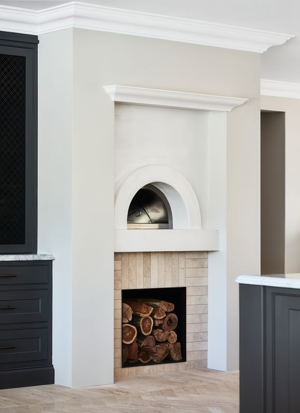 A refined traditional interior detail featuring a built-in fireplace with a pale stone or tile surround, arched firebox, and decorative white mantel and cornice mouldings. The fireplace is integrated between dark painted kitchen cabinetry with light stone benchtops. Cut firewood is stored in the open hearth, and pale timber flooring extends across the room.