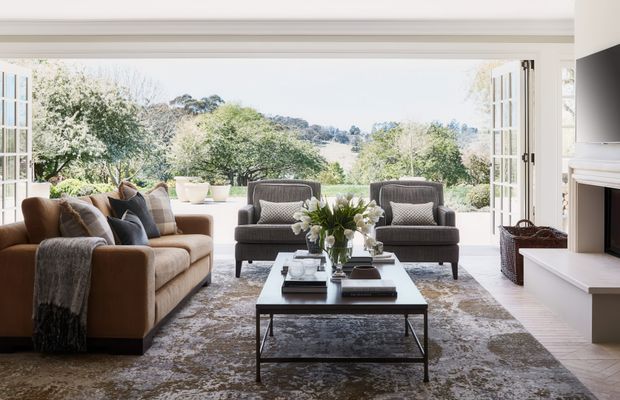 Bright, refined living room with a tan sofa, two dark upholstered armchairs, a rectangular glass-topped coffee table, and a large patterned rug. Full-height French doors open onto a leafy landscape, while a white fireplace and built-in shelving frame the right side. The room has a classic, neutral interior with abundant natural light and a strong indoor–outdoor connection.