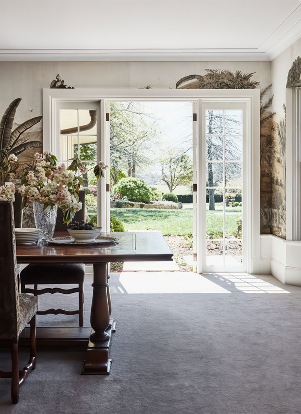 A bright, refined dining room with a traditional table and upholstered dining chairs positioned beside large white-framed glazed French doors. The room features pale grey carpet, white wainscoting and ceiling mouldings, warm neutral walls, botanical wallpaper or mural panels, and abundant natural light opening onto a formal landscaped garden. A floral arrangement and table setting add an elegant, lived-in character.