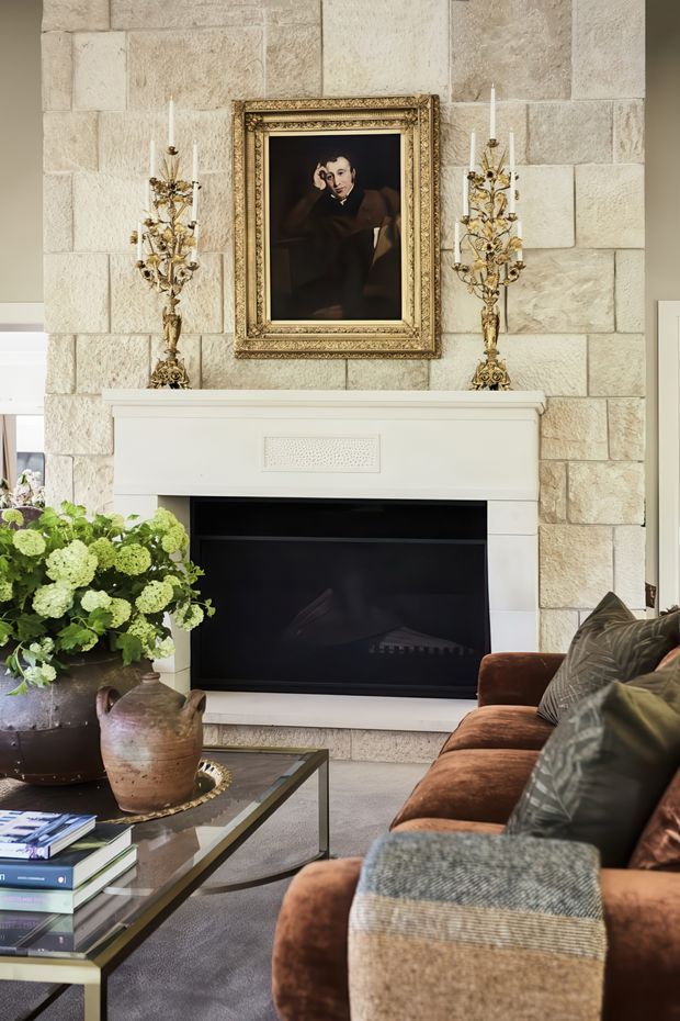 A refined living room centered on a traditional fireplace with a pale stone-block feature wall and white mantel. An ornate gilt-framed portrait hangs above the fireplace, flanked by decorative gold-toned candleholders. The room includes a deep brown velvet sectional sofa with textured throws, a glass-topped coffee table, and substantial potted greenery. The styling is warm, classic, and layered.