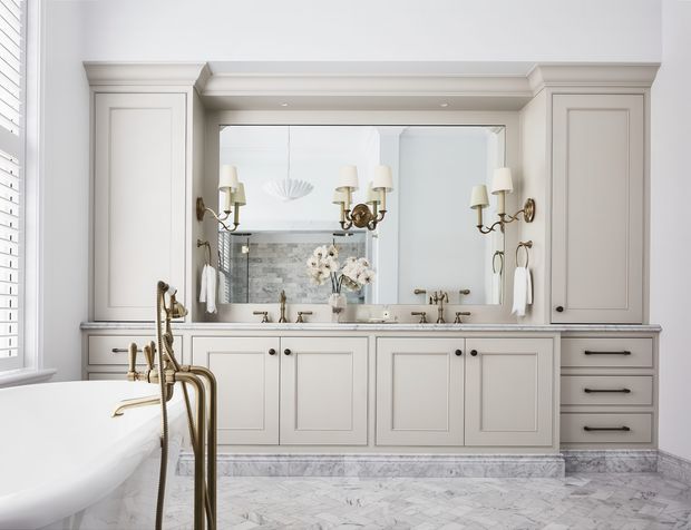 Elegant, bright traditional bathroom featuring a freestanding white bathtub, extensive pale painted vanity cabinetry, a large framed mirror, brass wall sconces and tapware, and a light marble-patterned floor. Shutters at the window and detailed cornice and panel mouldings reinforce the refined classic character.